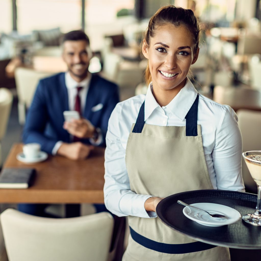 Portrait of happy waitress holding a tray with a lemon tart with raspberry sorbe and looking at camera. There is a businessman sitting at the table in the background.